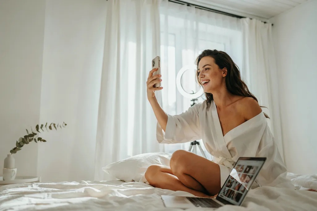 A woman in a white robe photographing herself on a bright white bed, ring light and laptop beside her