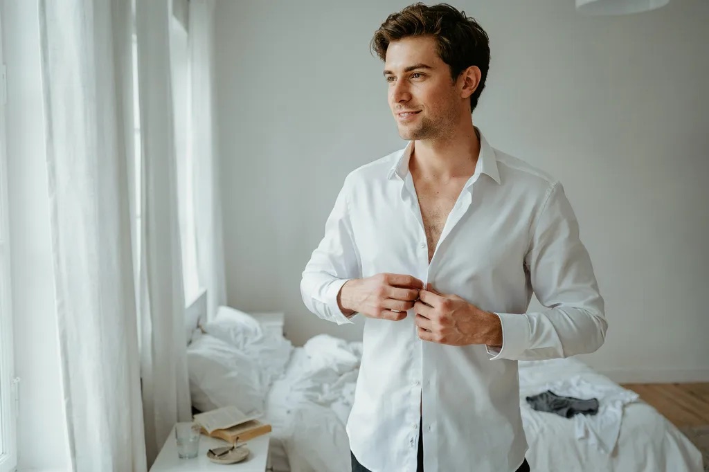 A man buttoning a white shirt in a sunlit bedroom