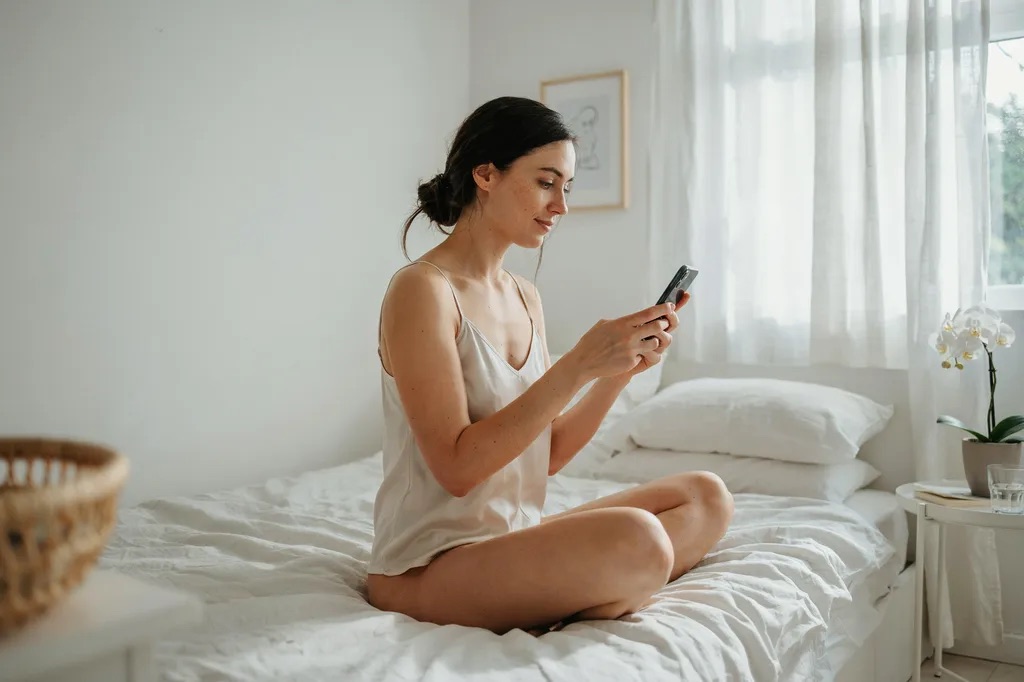 A woman in a white camisole checking her phone on the edge of the bed